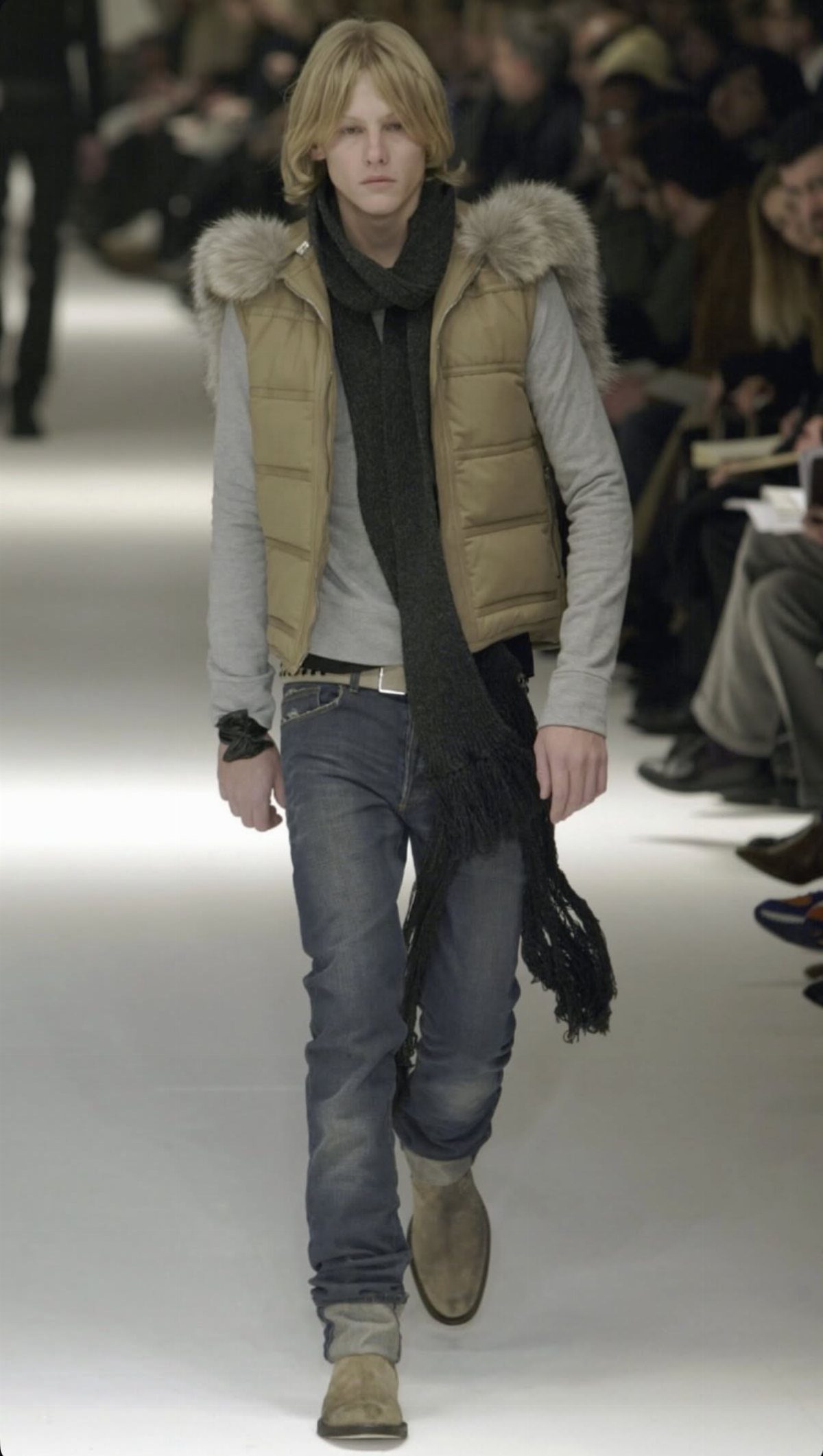 Blonde man walking a fashion runway in a tan fur-trimmed vest and blue jeans.
