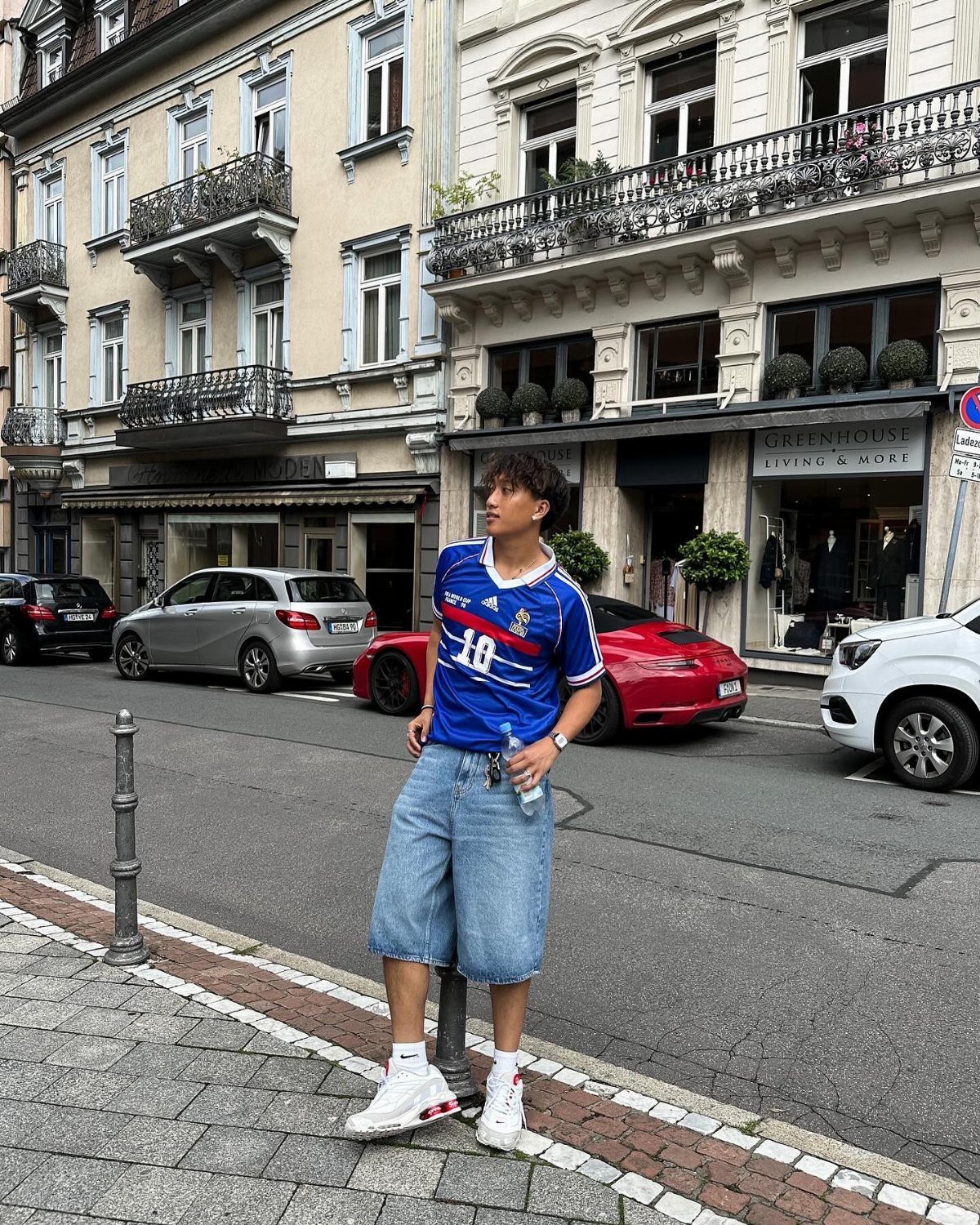 A young man posing in a blue football jersey and denim shorts on a city street.