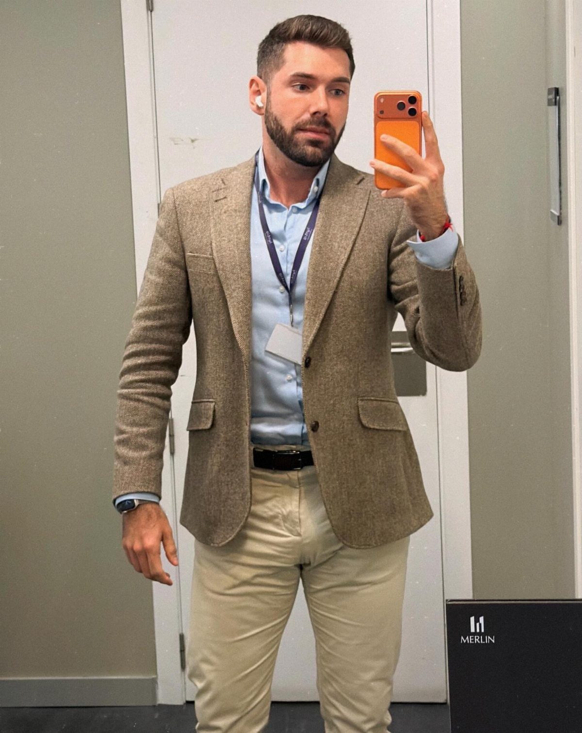 A man posing in a tan blazer, blue shirt, and beige trousers for a mirror selfie.