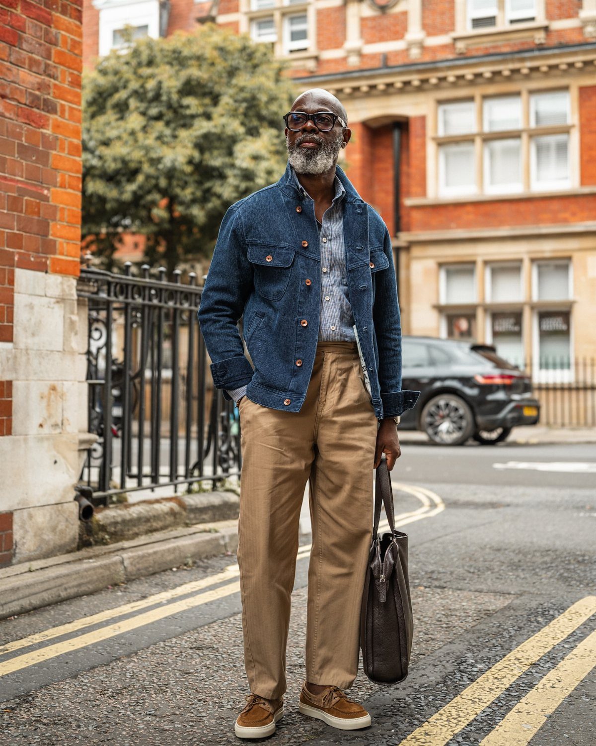 A stylish older man wearing a denim jacket and tan trousers standing on a city street.