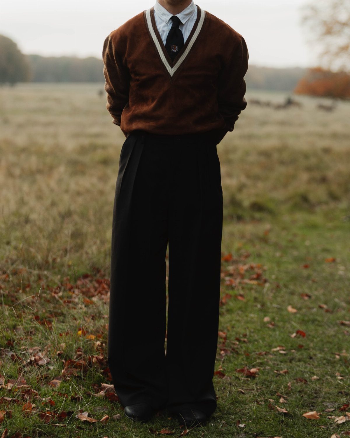 A person wearing a brown sweater, white shirt, tie, and black trousers standing in a field.