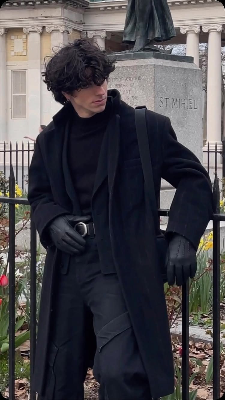 A man in an all-black layered outfit standing by a black metal fence outdoors.