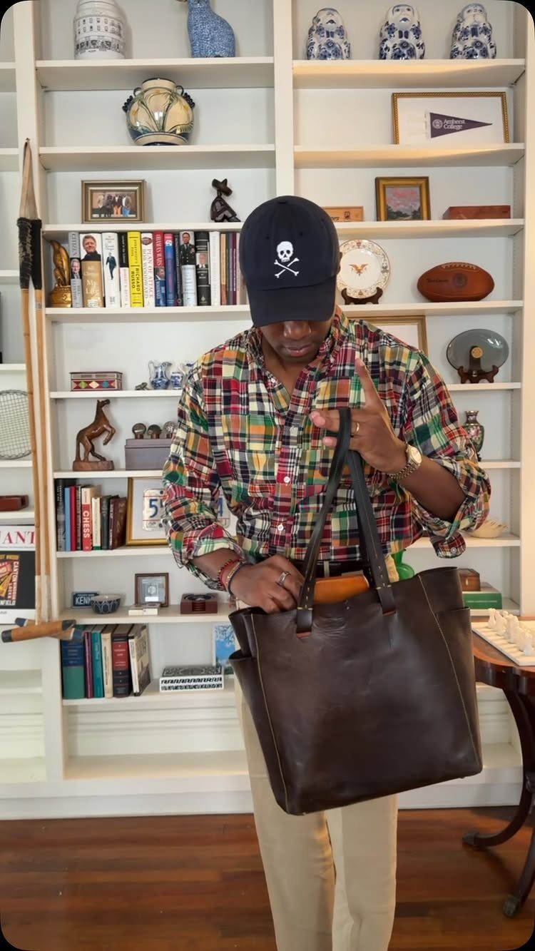 Man wearing a colorful Madras plaid shirt and a skull cap holding a leather bag.