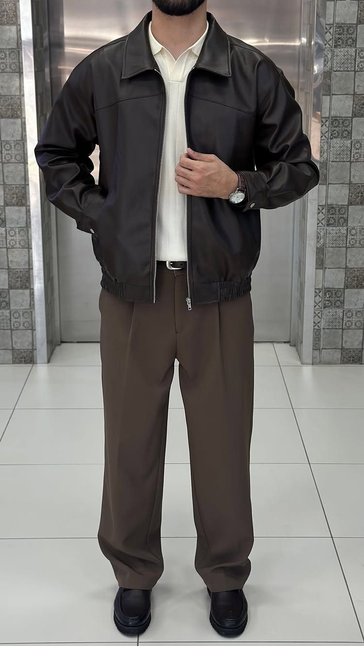 Man wearing a brown leather jacket, cream polo, and brown pleated trousers with black loafers.
