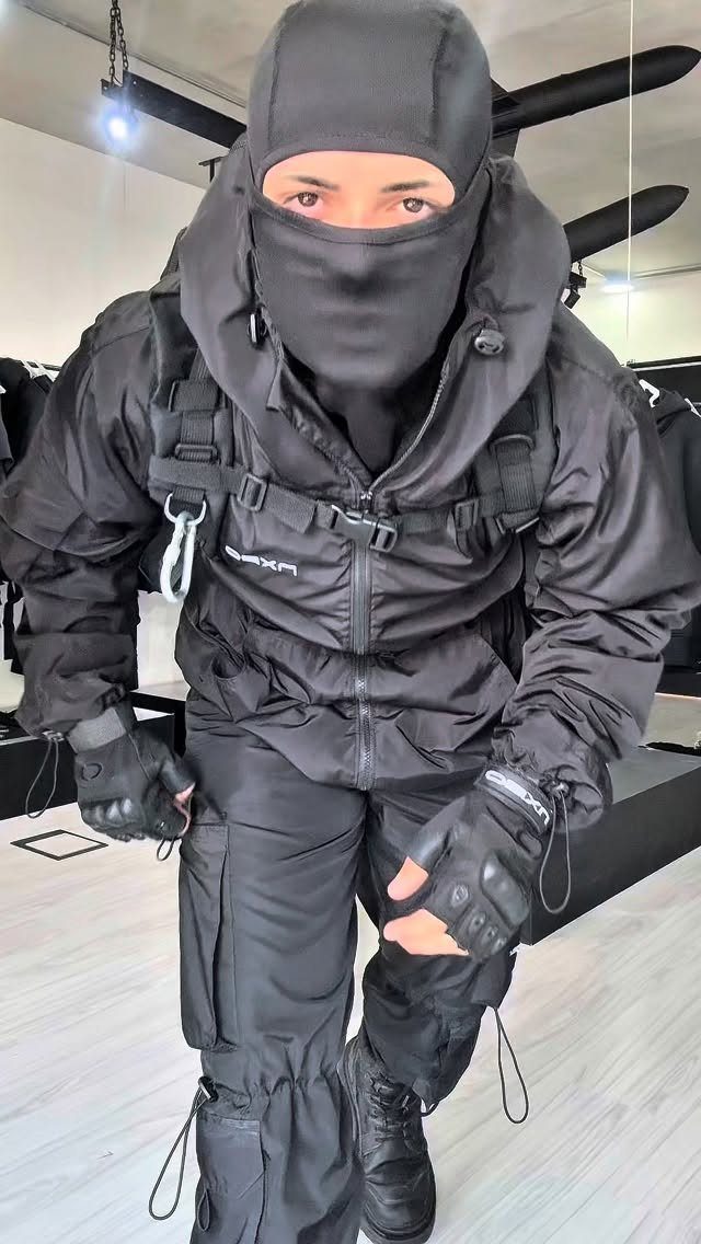 A person dressed in full black tactical techwear including a balaclava, hooded jacket, and cargo pants.