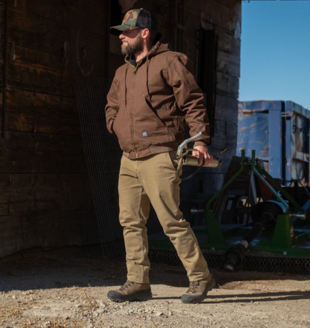 Man in brown work jacket and tan trousers standing outdoors near a wooden building.