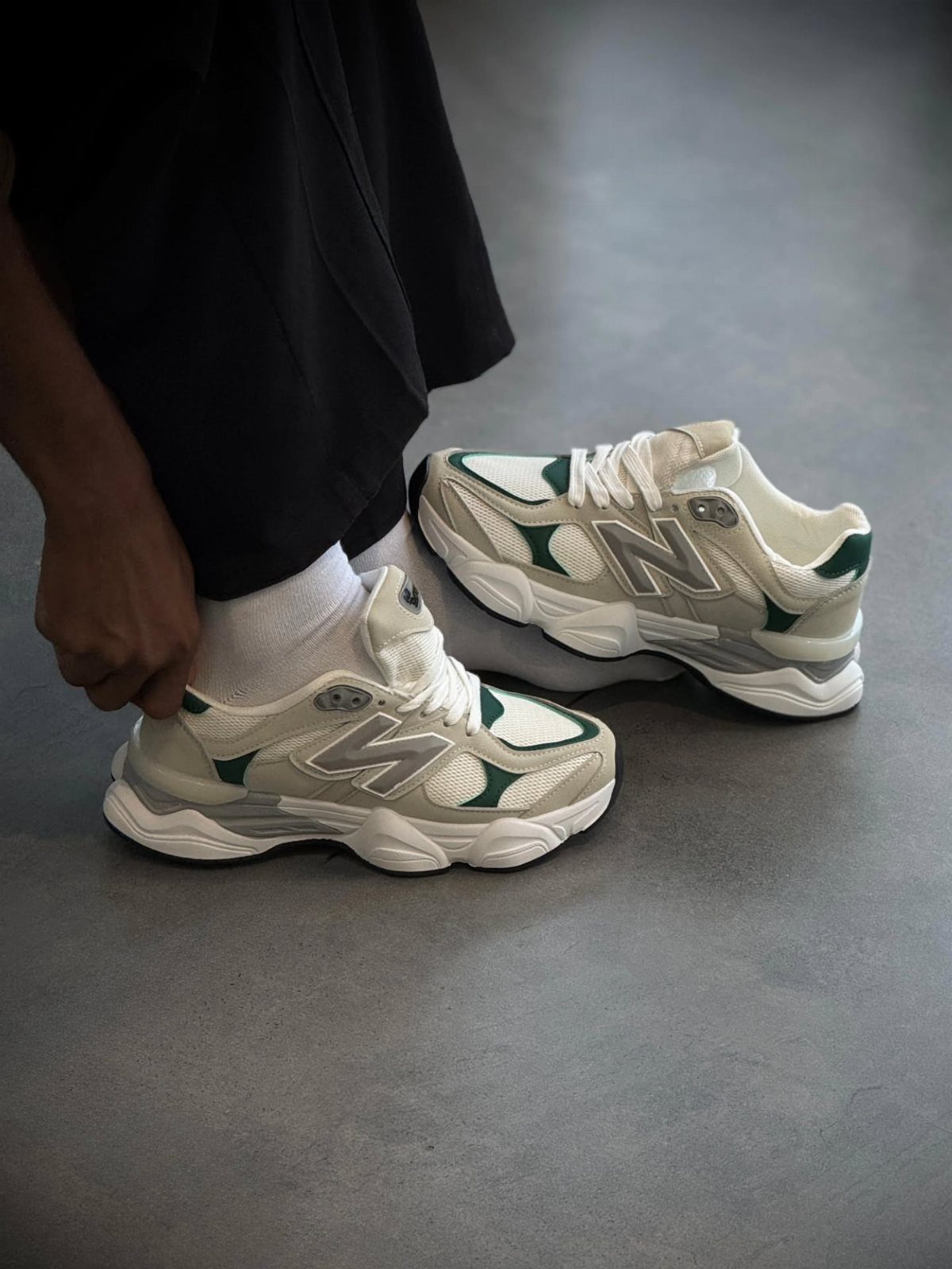 A person wearing cream and green chunky New Balance sneakers with white socks on concrete.
