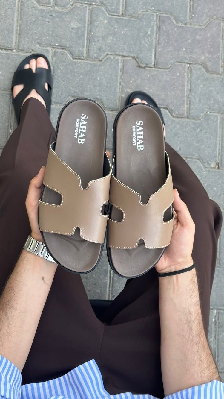 A person holding a pair of tan leather slide sandals over brown trousers.