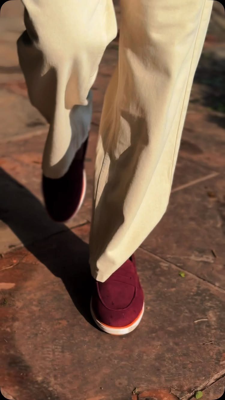 Burgundy loafers worn with cream trousers walking on a stone pavement in sunlight.