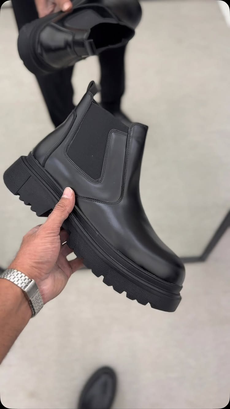 A hand holding a black leather Chelsea boot with a chunky lugged sole.