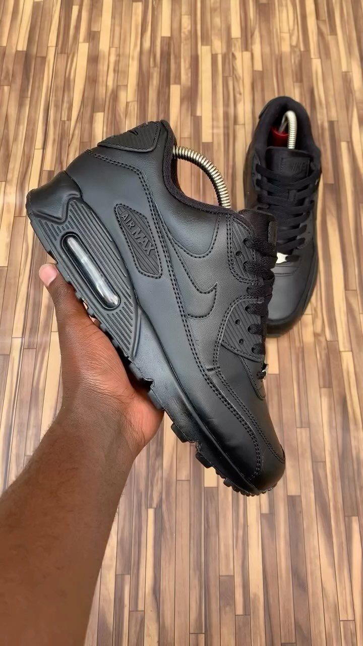 A hand holding a black Nike Air Max sneaker over a wooden floor.