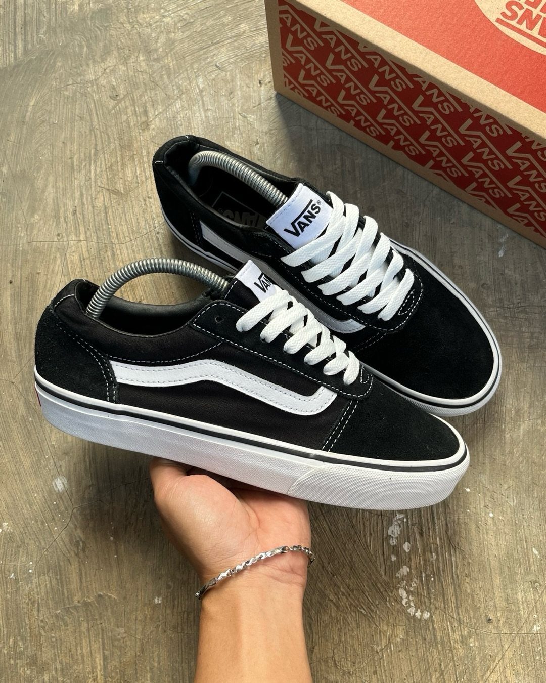 A hand holding a pair of black and white Vans Ward sneakers against a concrete background.