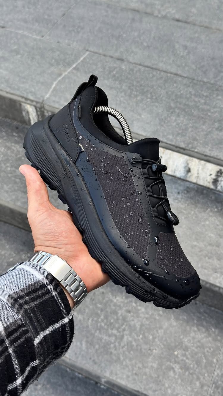 A hand holding a black sneaker covered in water droplets against a stone background.