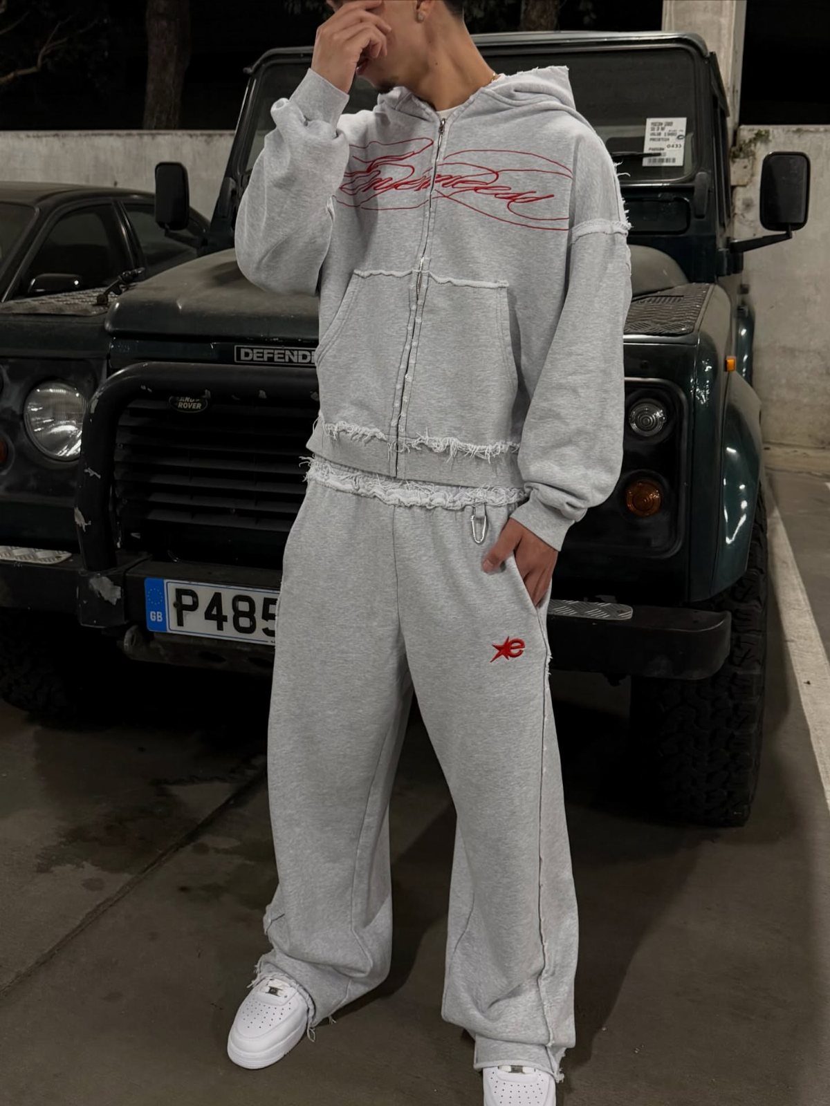 A man wearing a grey two-piece fleece tracksuit and white sneakers standing by a dark vehicle.