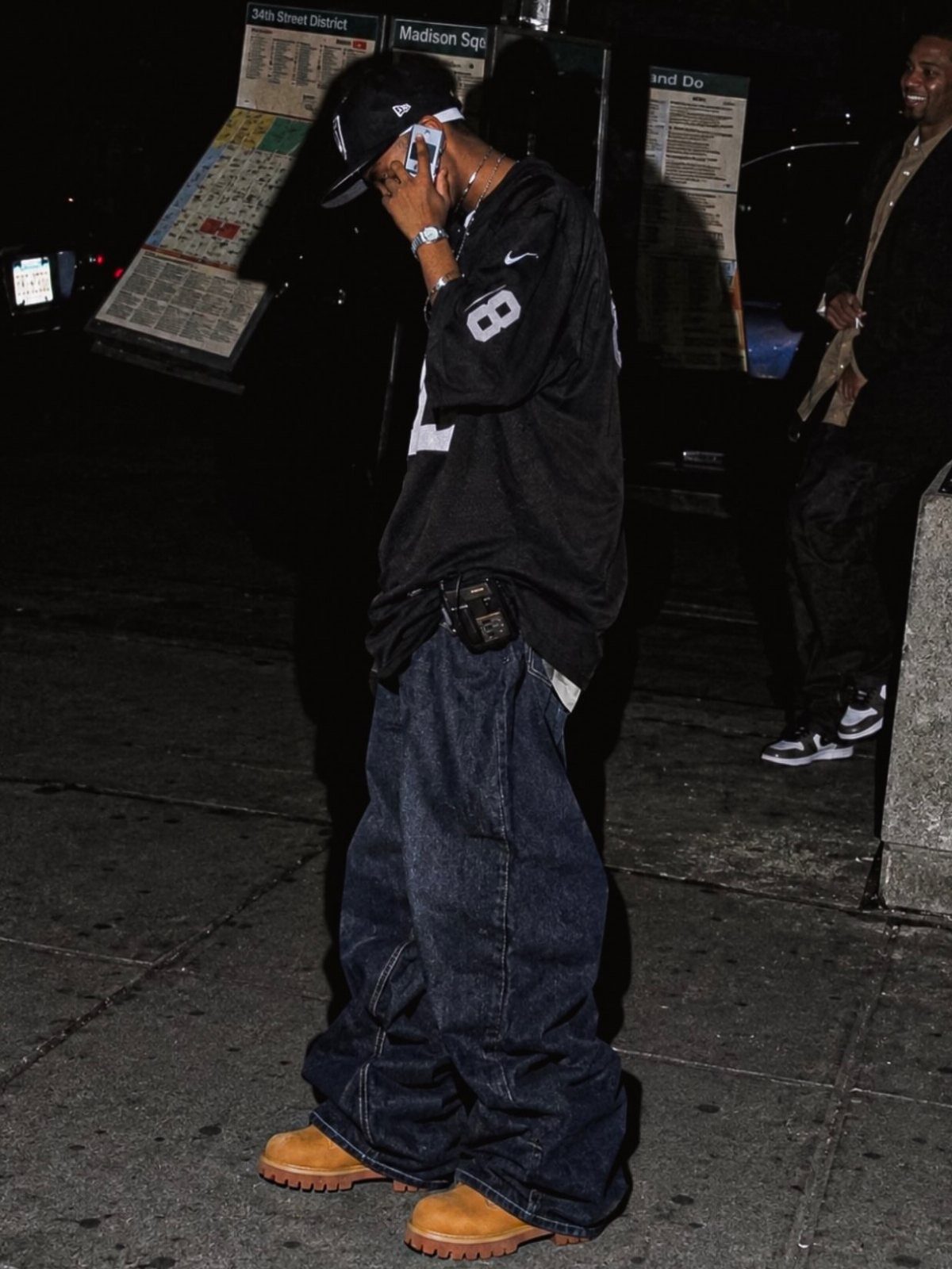 A man wearing baggy dark denim jeans, a black shirt, and tan boots on a city sidewalk.