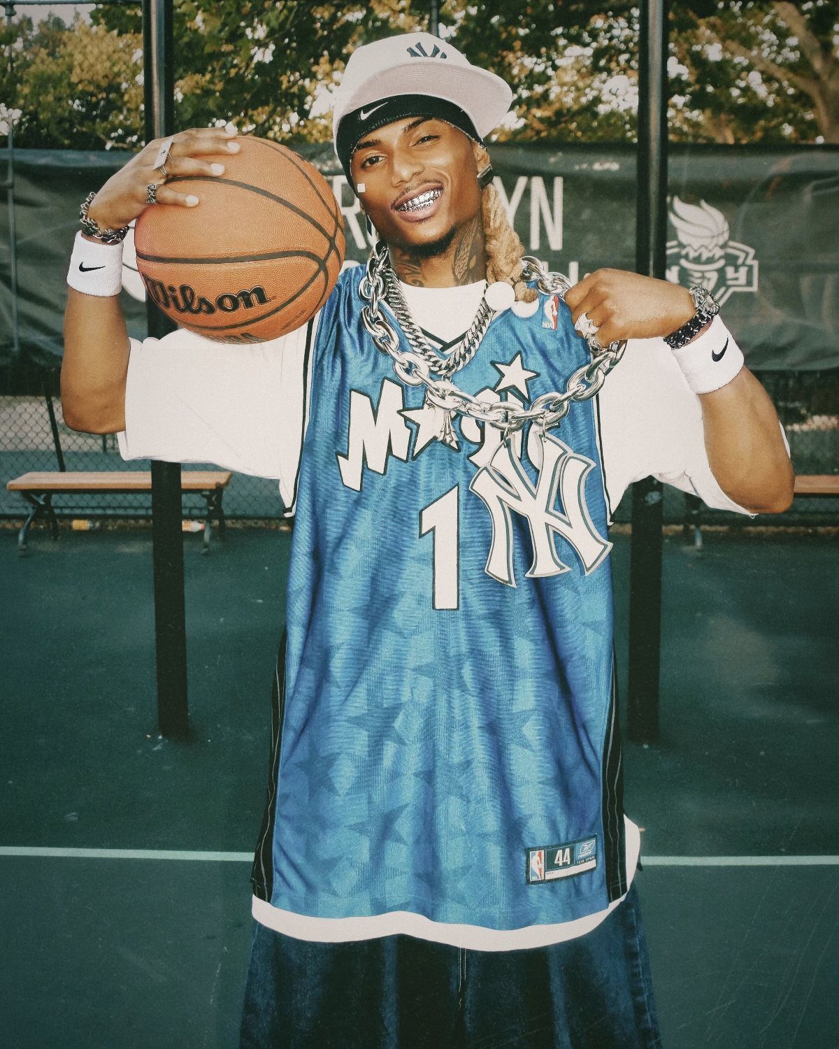 A man in a blue basketball jersey and baggy jeans holding a basketball outdoors.