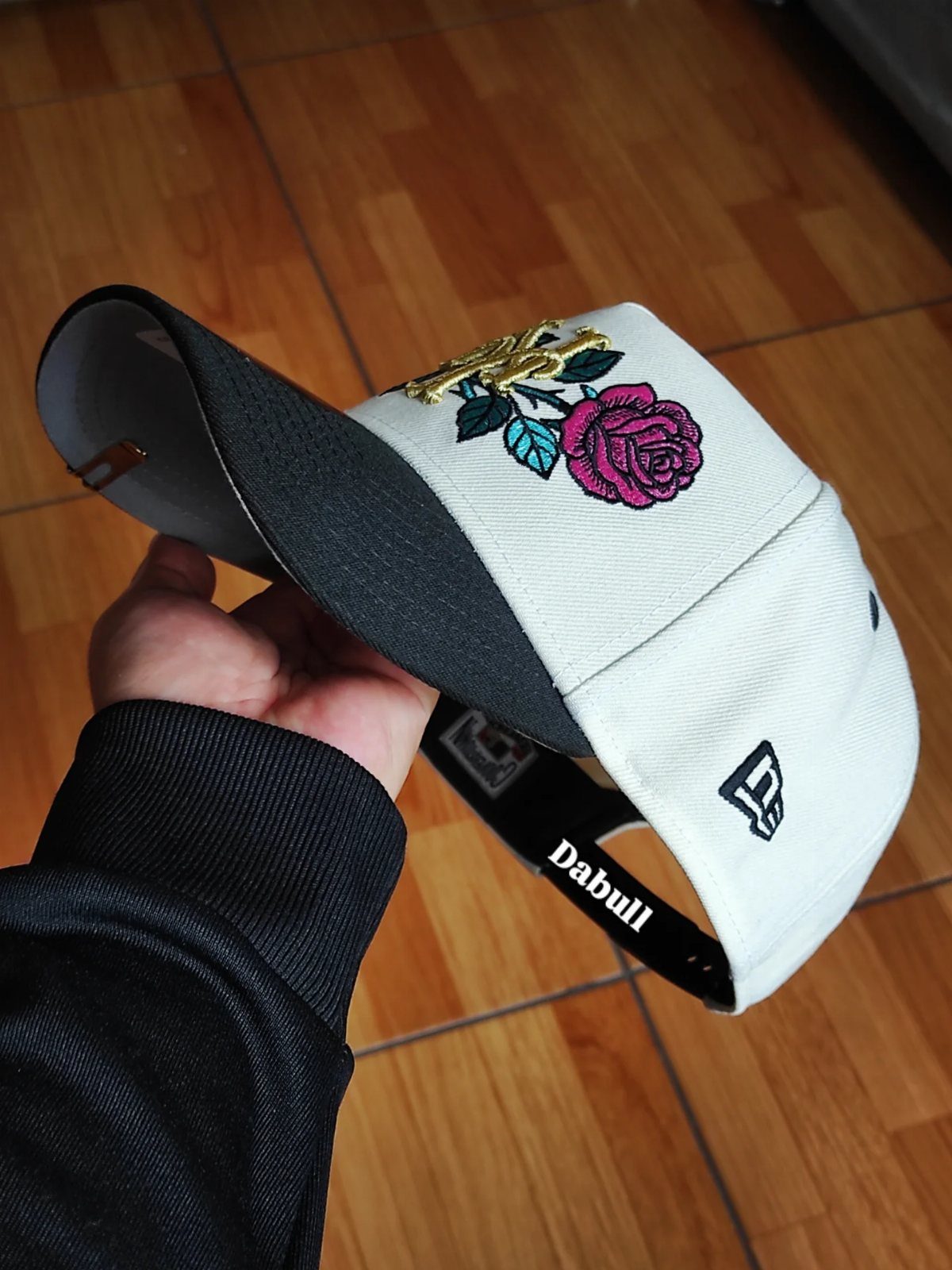 A hand holding a white and black baseball cap with pink rose embroidery.