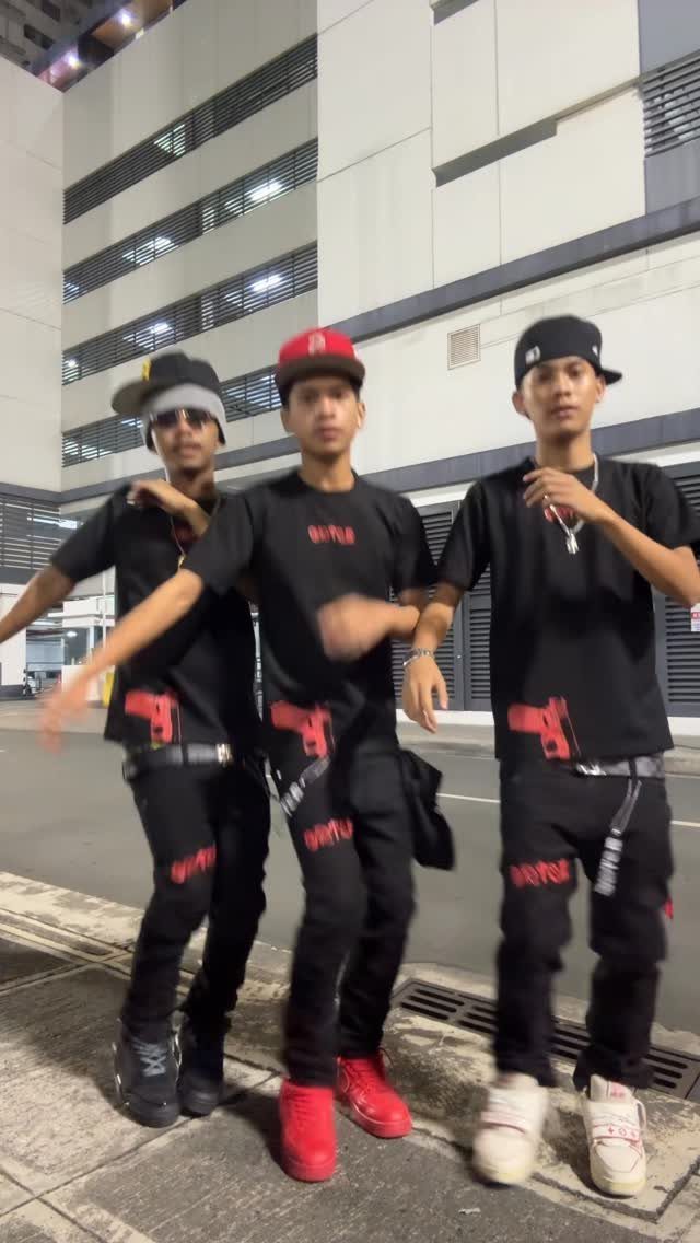 Three young men posing in black graphic t-shirts, black pants, and various caps at night.