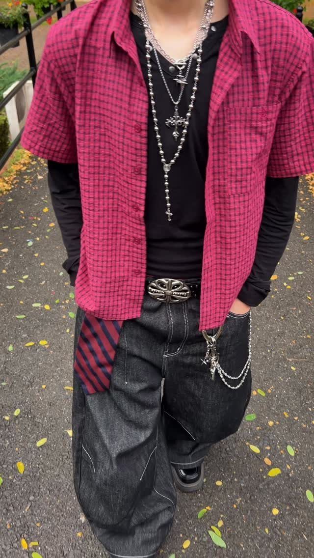 Person wearing a pink checkered shirt, black long sleeves, and baggy black denim jeans with silver accessories.