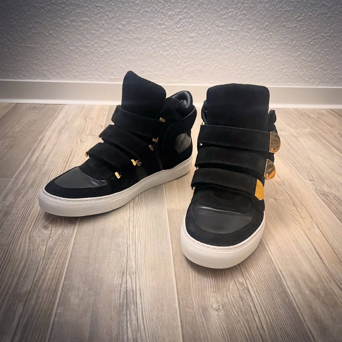 Black high-top sneakers with wide velcro straps resting on a light wooden floor.