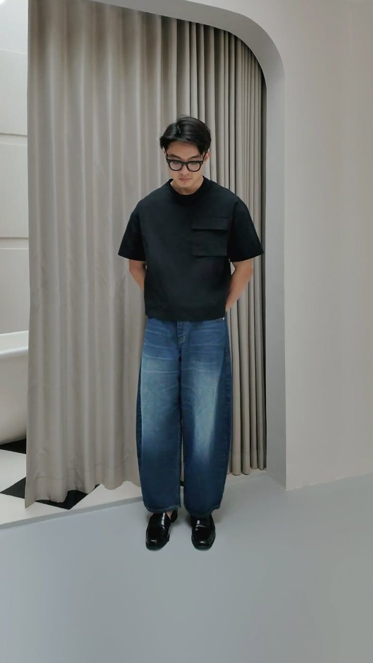 A man wearing black t-shirt and wide blue barrel jeans standing before grey curtains.