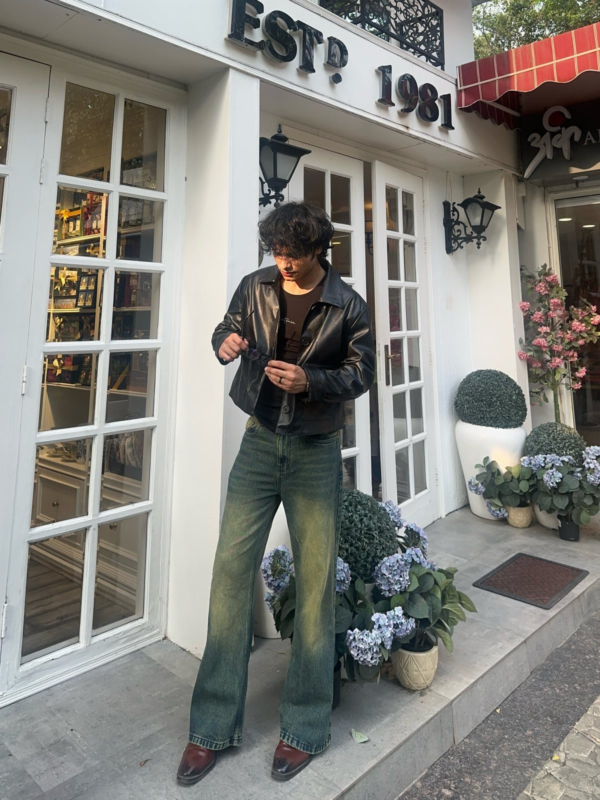 A man wearing dark green bootcut jeans and a black leather jacket standing outside a shop.