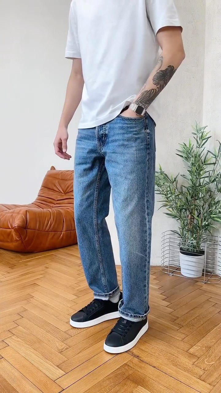 A man wearing blue denim jeans, a white t-shirt, and black sneakers standing indoors.
