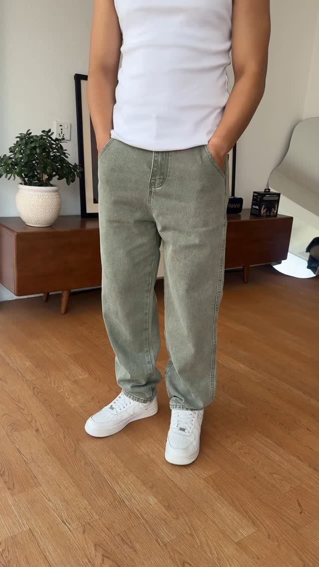 A person wearing olive green relaxed fit trousers and white sneakers standing on a wooden floor.
