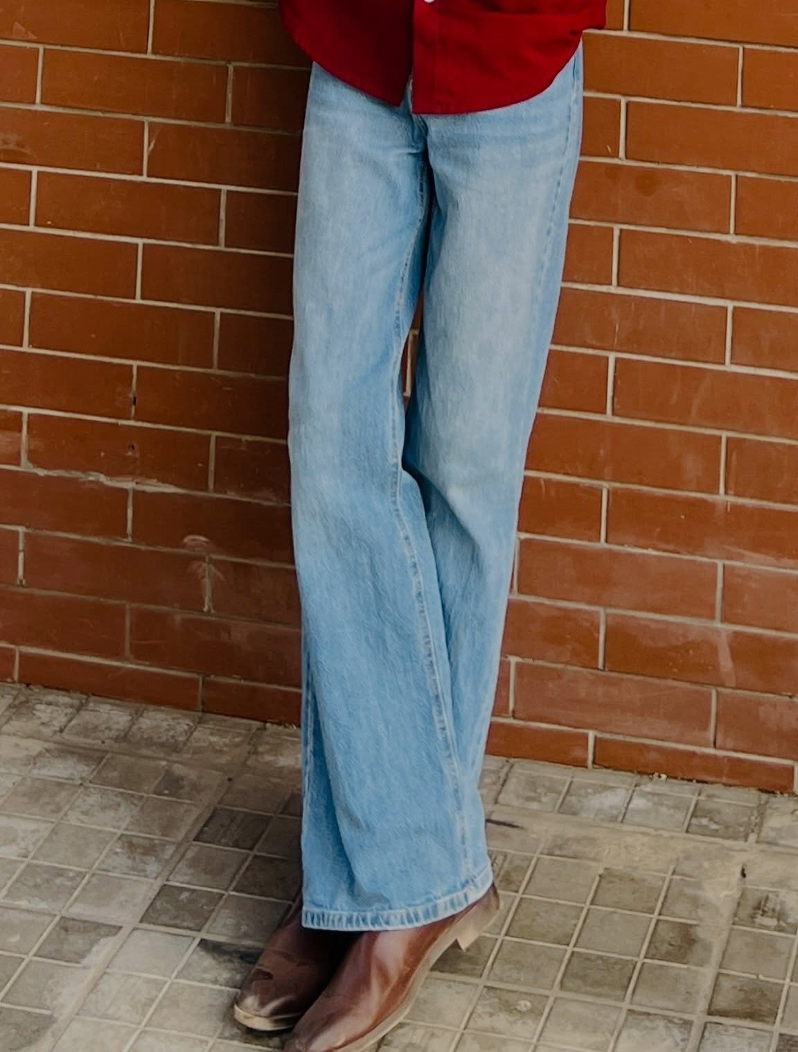 A person wearing light blue straight-leg jeans and brown leather boots against a brick wall.