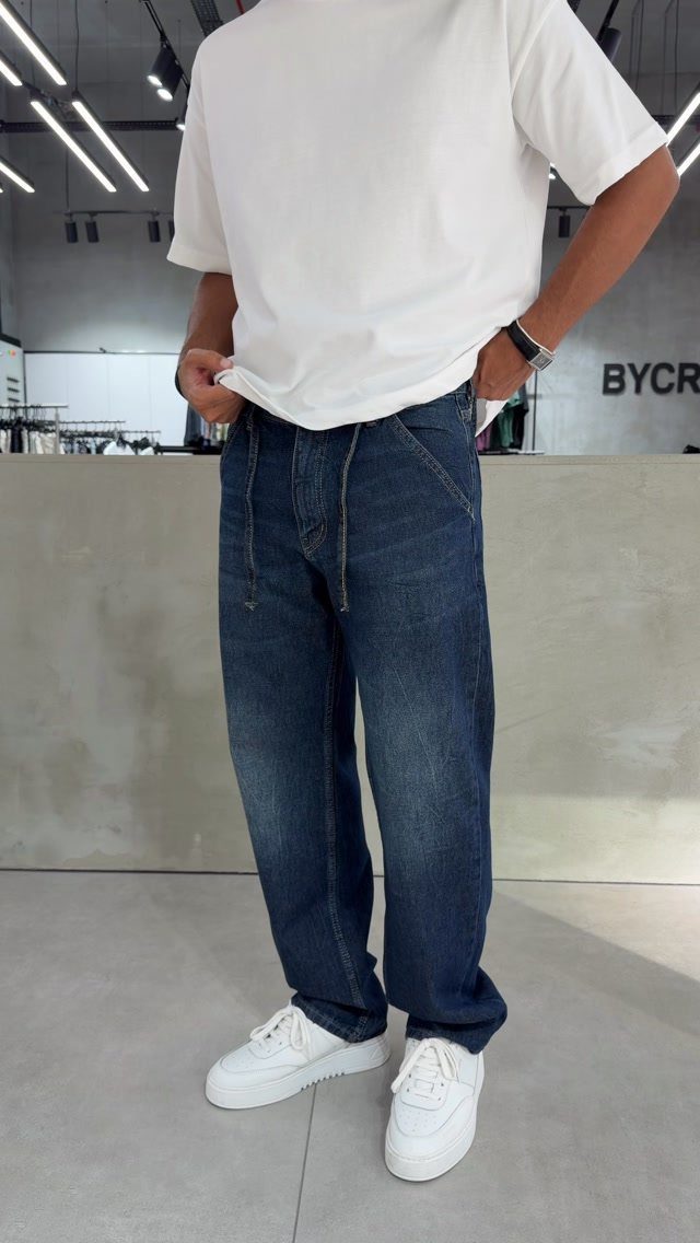 A person wearing wide-leg dark blue denim jeans and white sneakers standing indoors.