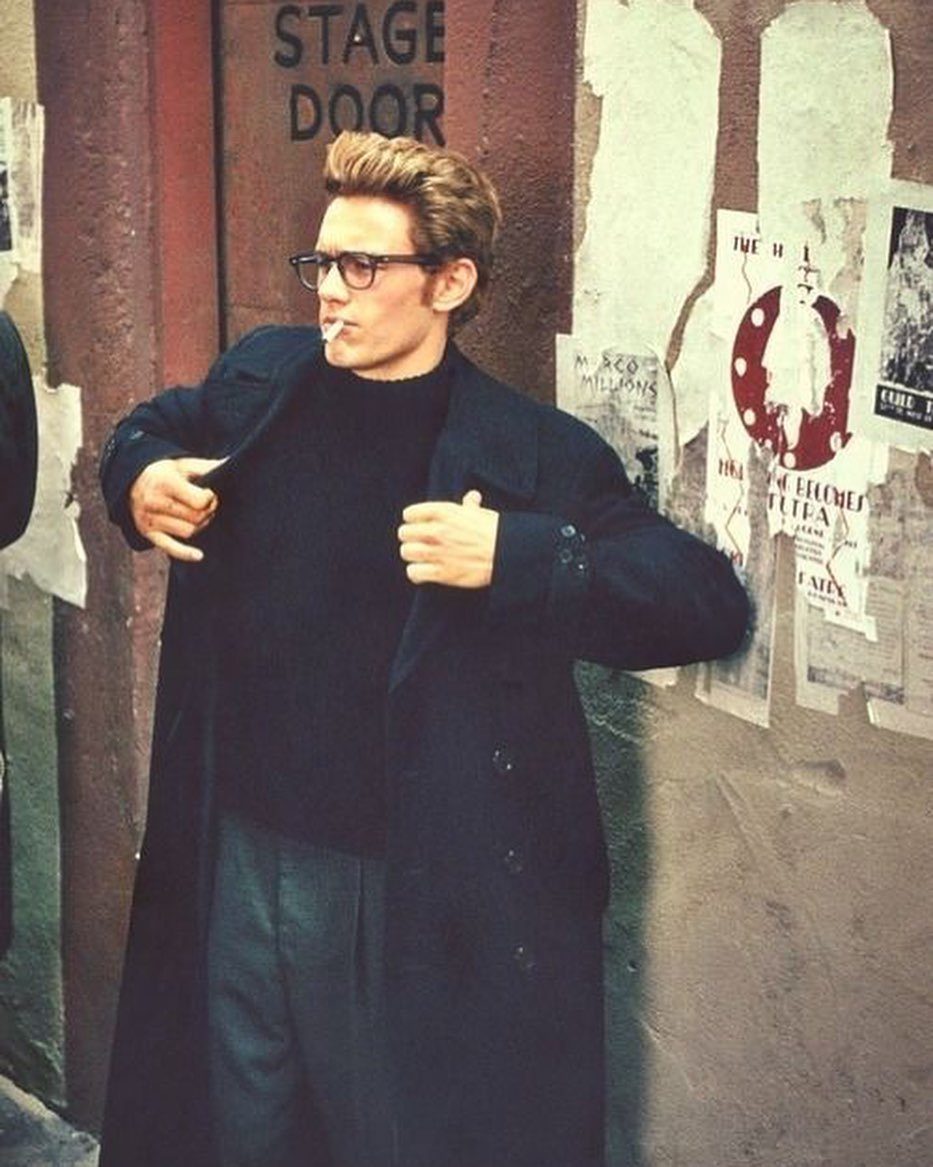 A stylish man in a black overcoat and turtleneck stands by a poster-covered wall.