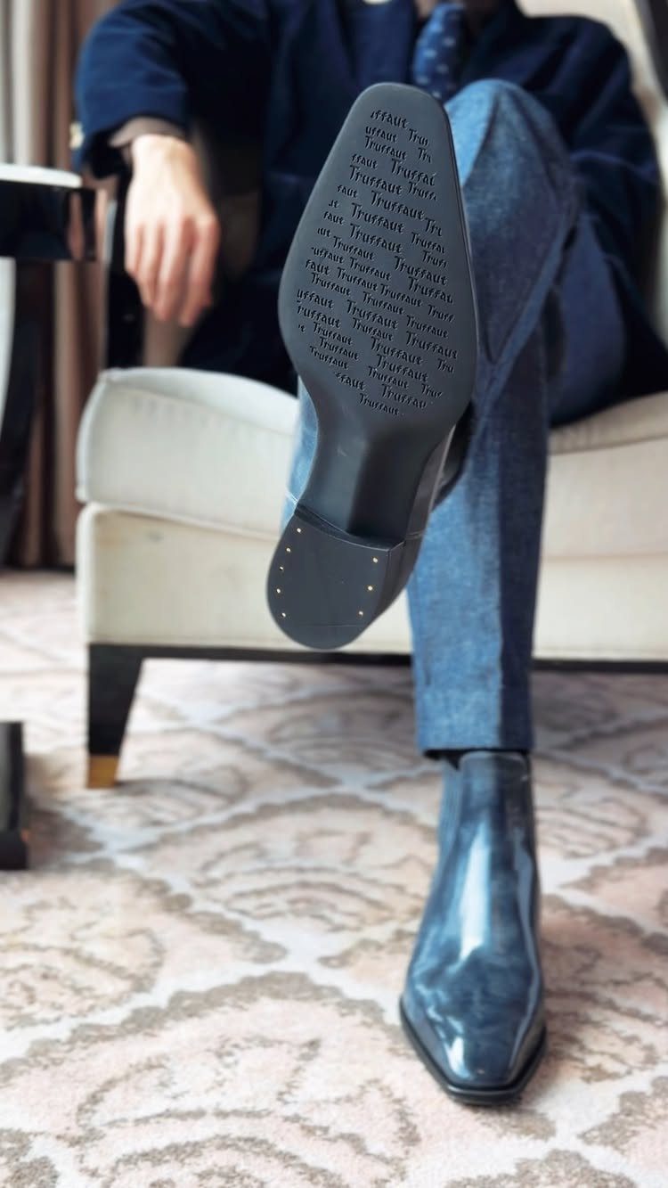 Polished blue leather Chelsea boots worn by a person sitting in a white armchair.