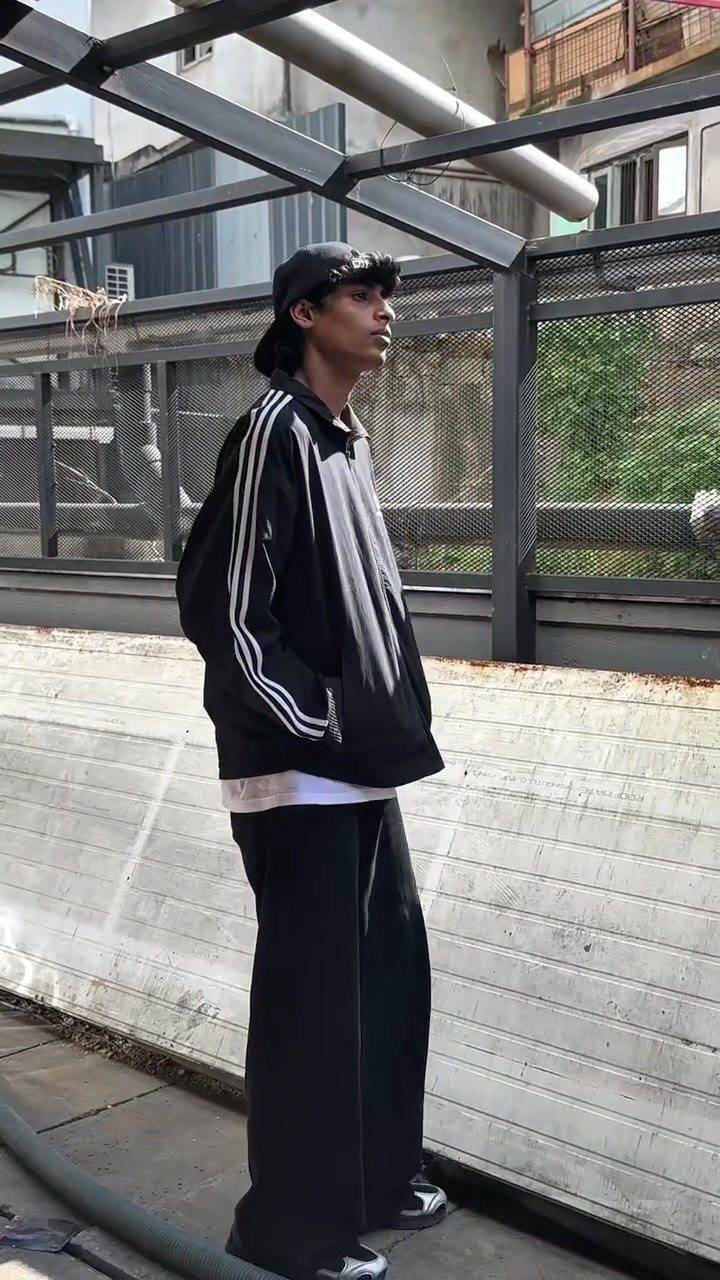 A young man wearing a black track jacket, wide-leg pants, and a black cap.