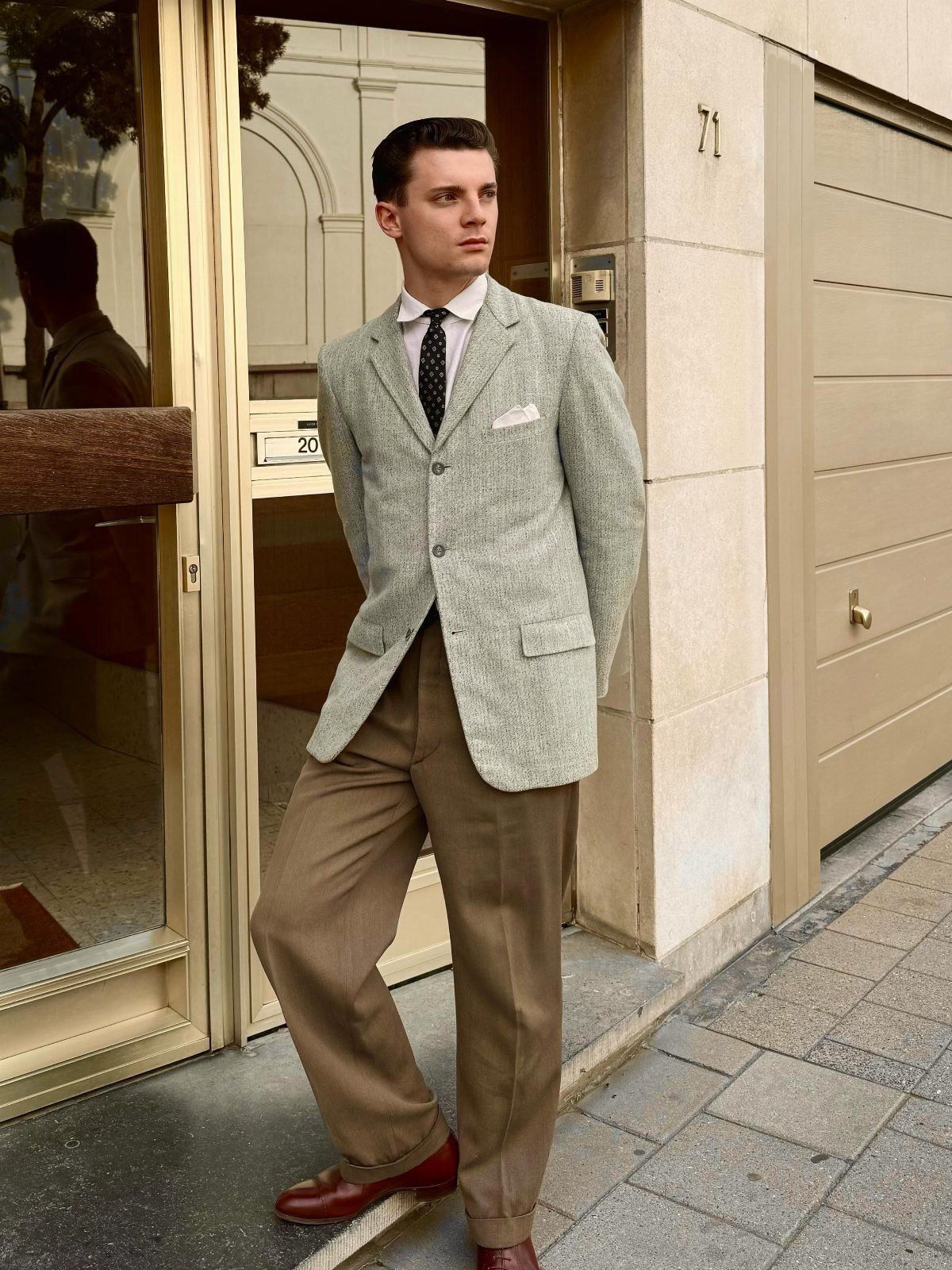 A man posing in a vintage-style grey blazer, tie, tan trousers, and leather shoes.