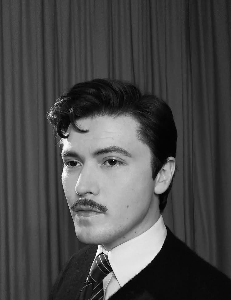 A young man with a vintage hairstyle and mustache wearing a striped necktie.