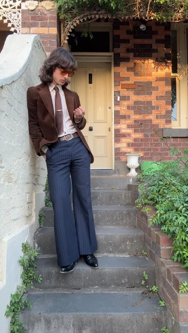 A man in a brown blazer and blue pinstriped wide-leg trousers standing on outdoor steps.