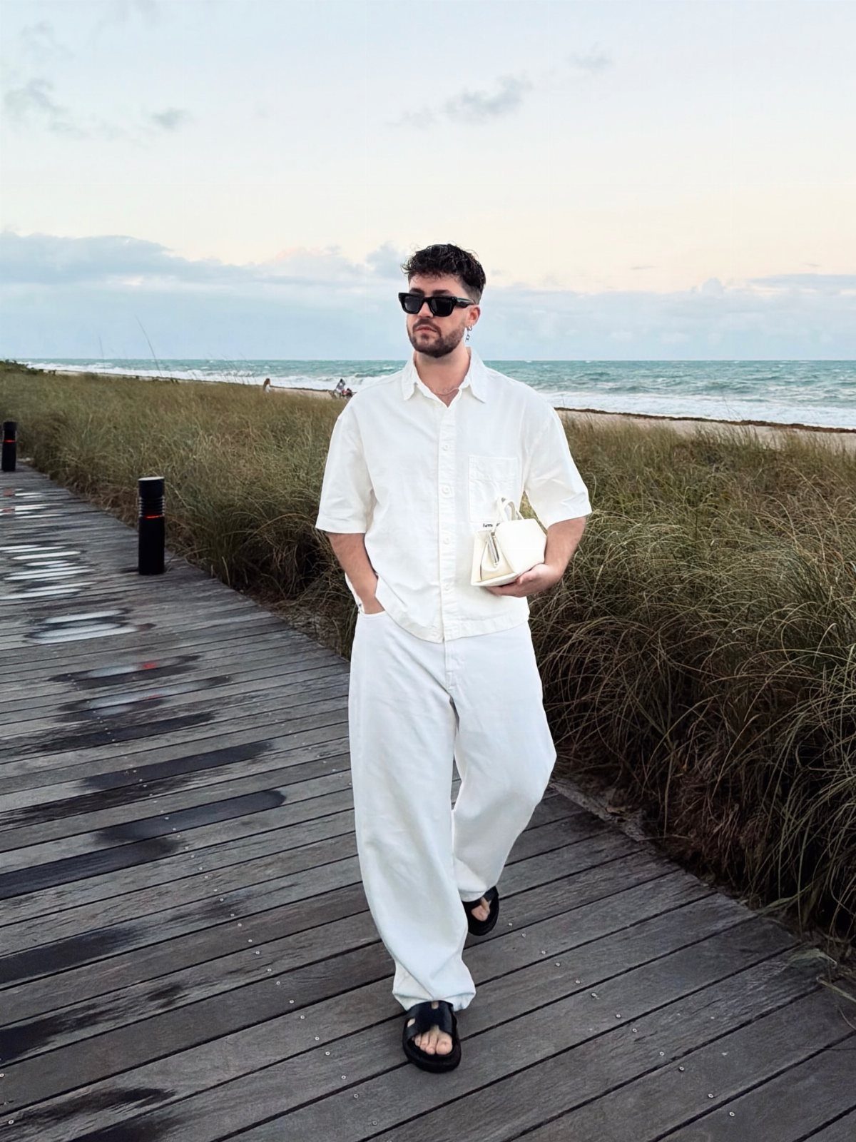 A man in an all-white linen outfit walking on a wooden boardwalk by the ocean.