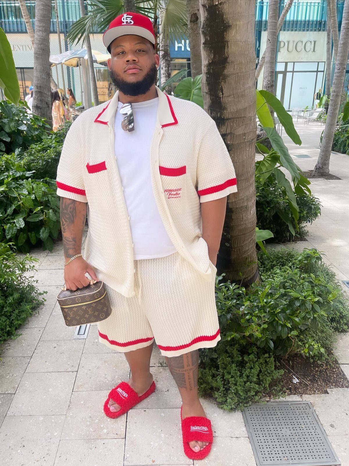 A man posing in a cream knit summer co-ord set with red accents and red slides.