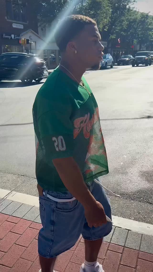 A young man wearing a green mesh jersey and denim shorts walking on a sunny city street.