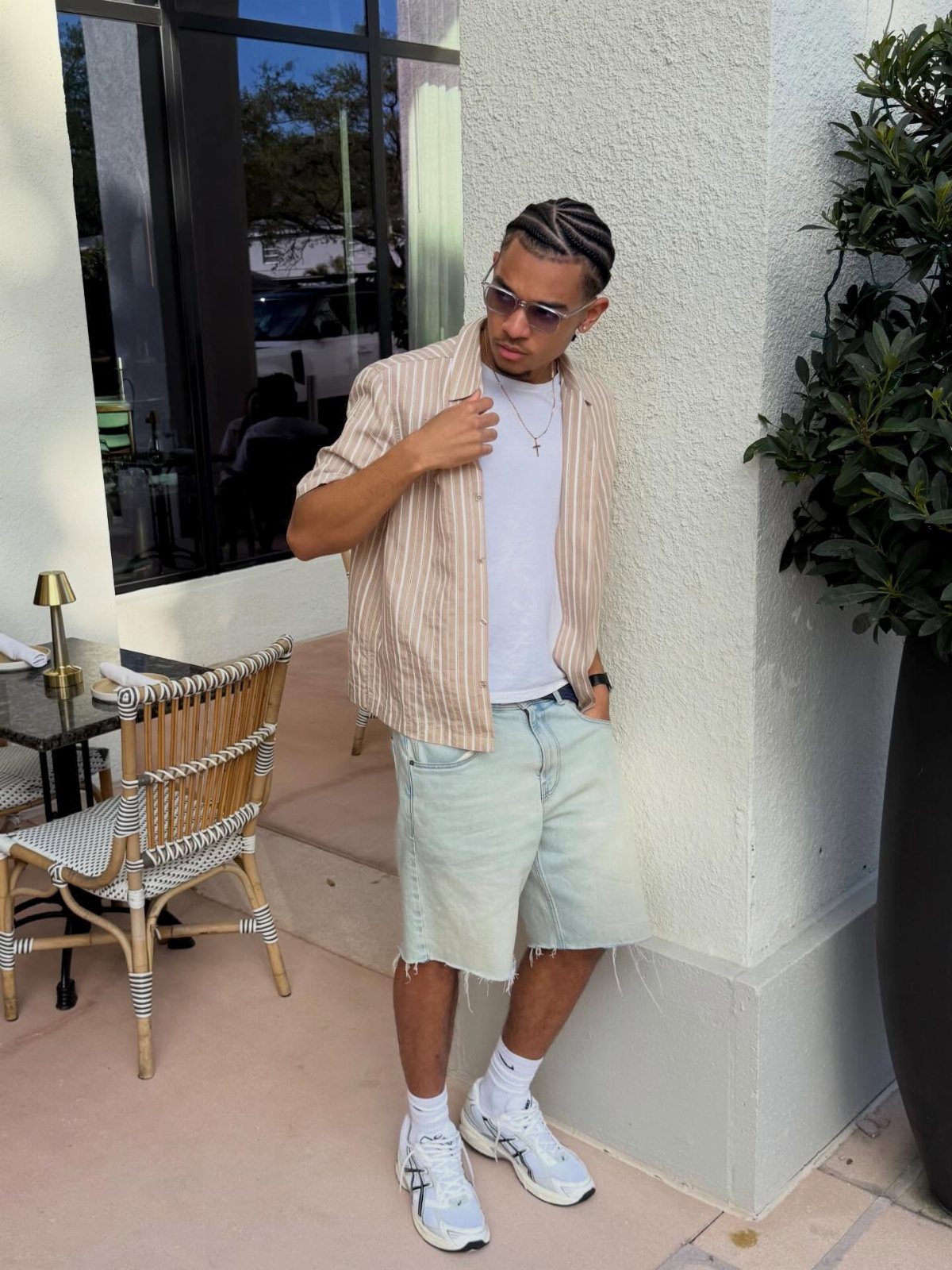 A man in a striped shirt and denim shorts posing on a sunny sidewalk.