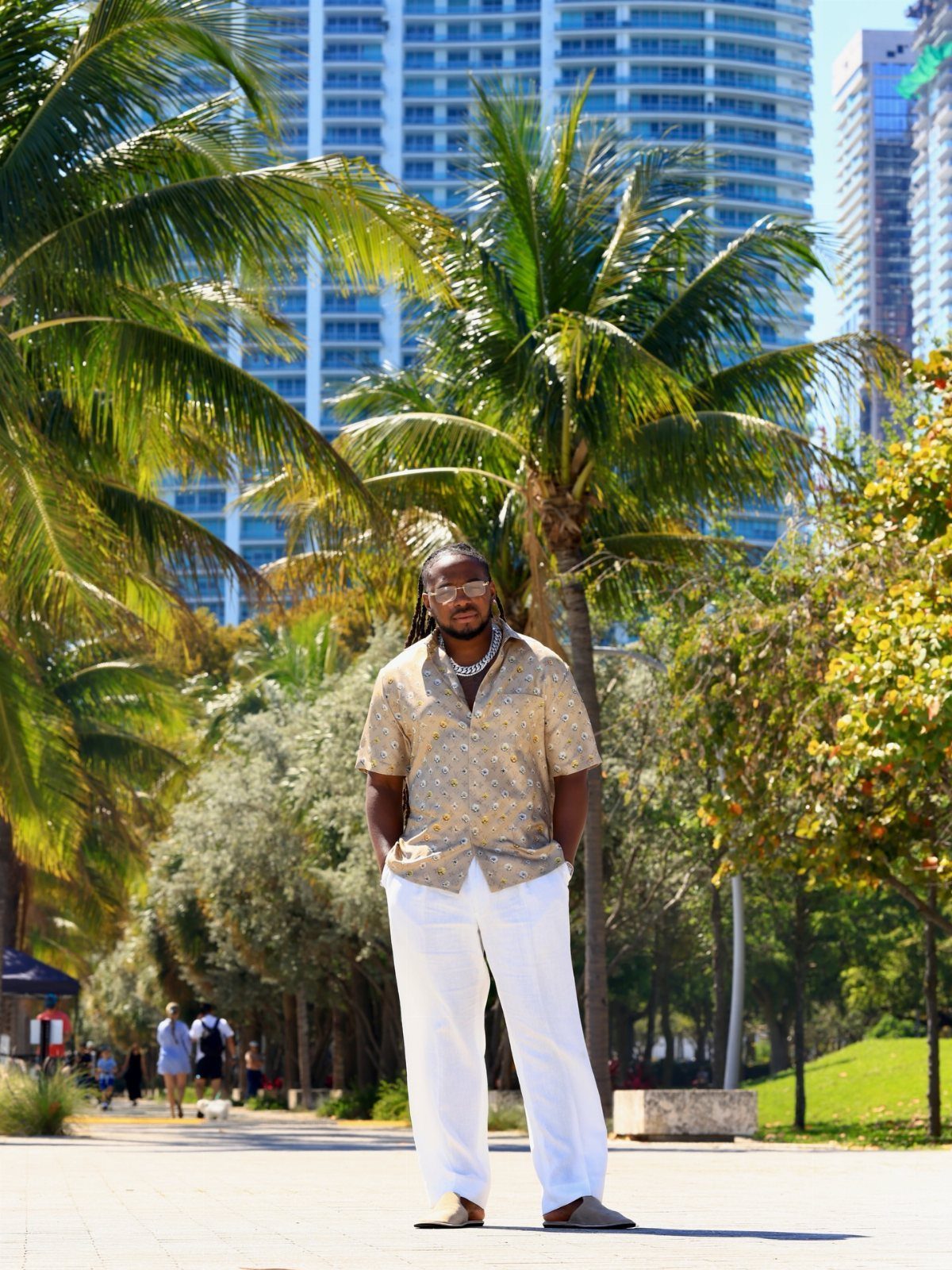 A man in a patterned shirt and white trousers standing among palm trees in Miami.