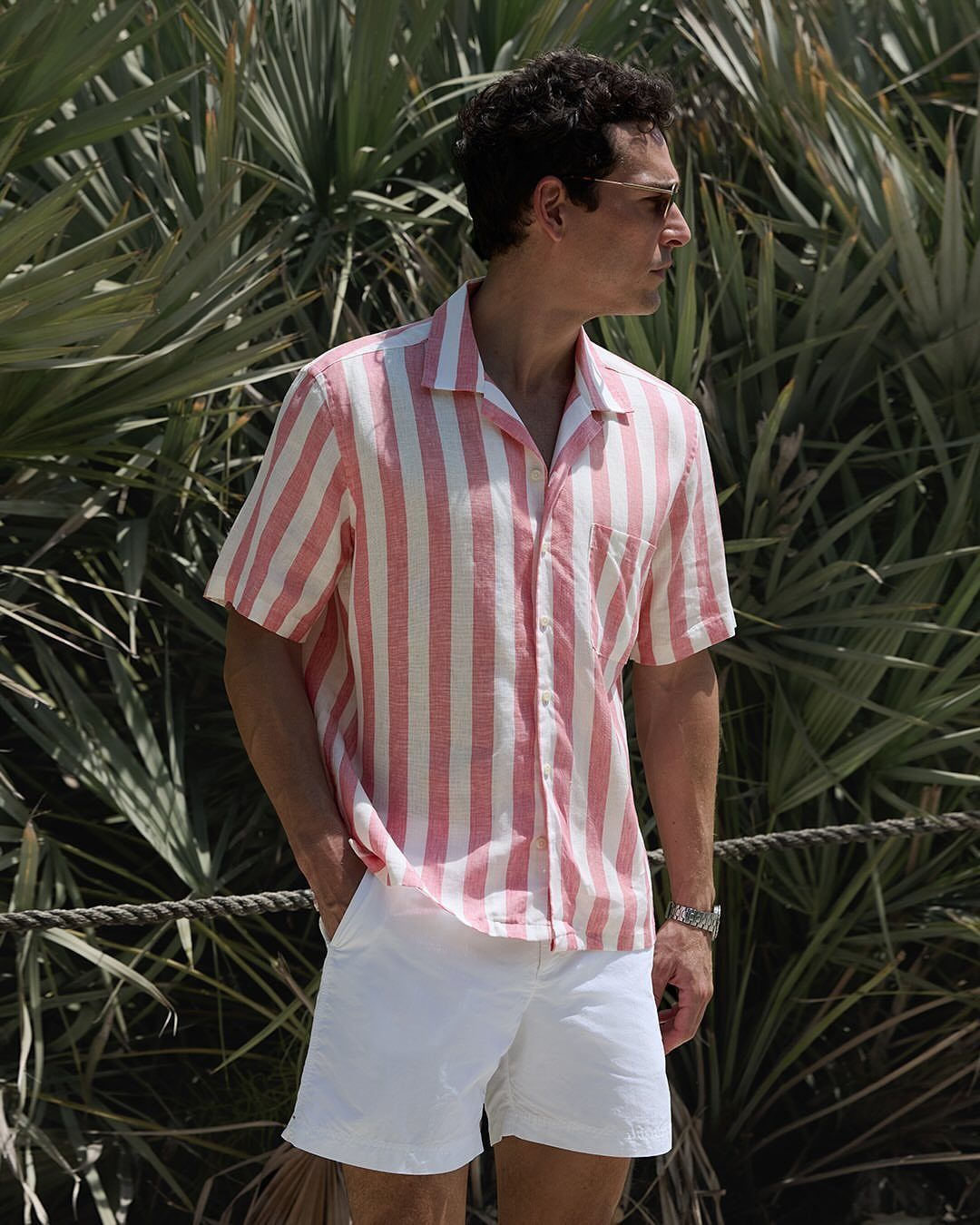 A man wearing a striped linen shirt and white shorts standing in front of palm trees.