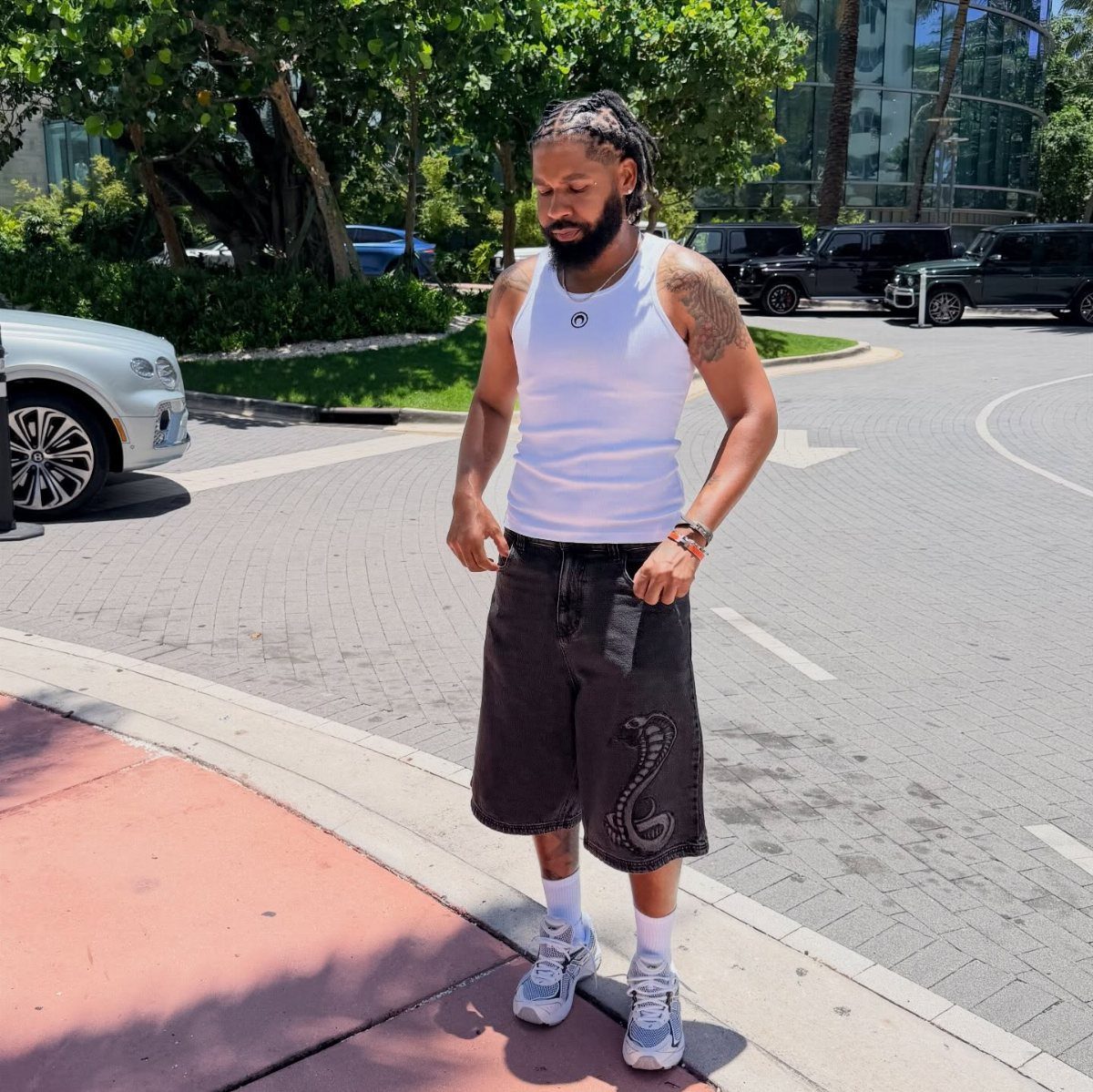 A man in a white tank top and black shorts standing on a sunny city street.