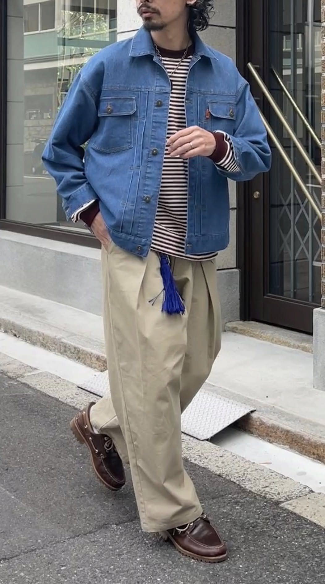 A man wearing a blue denim jacket, striped shirt, and beige trousers standing on a sidewalk.