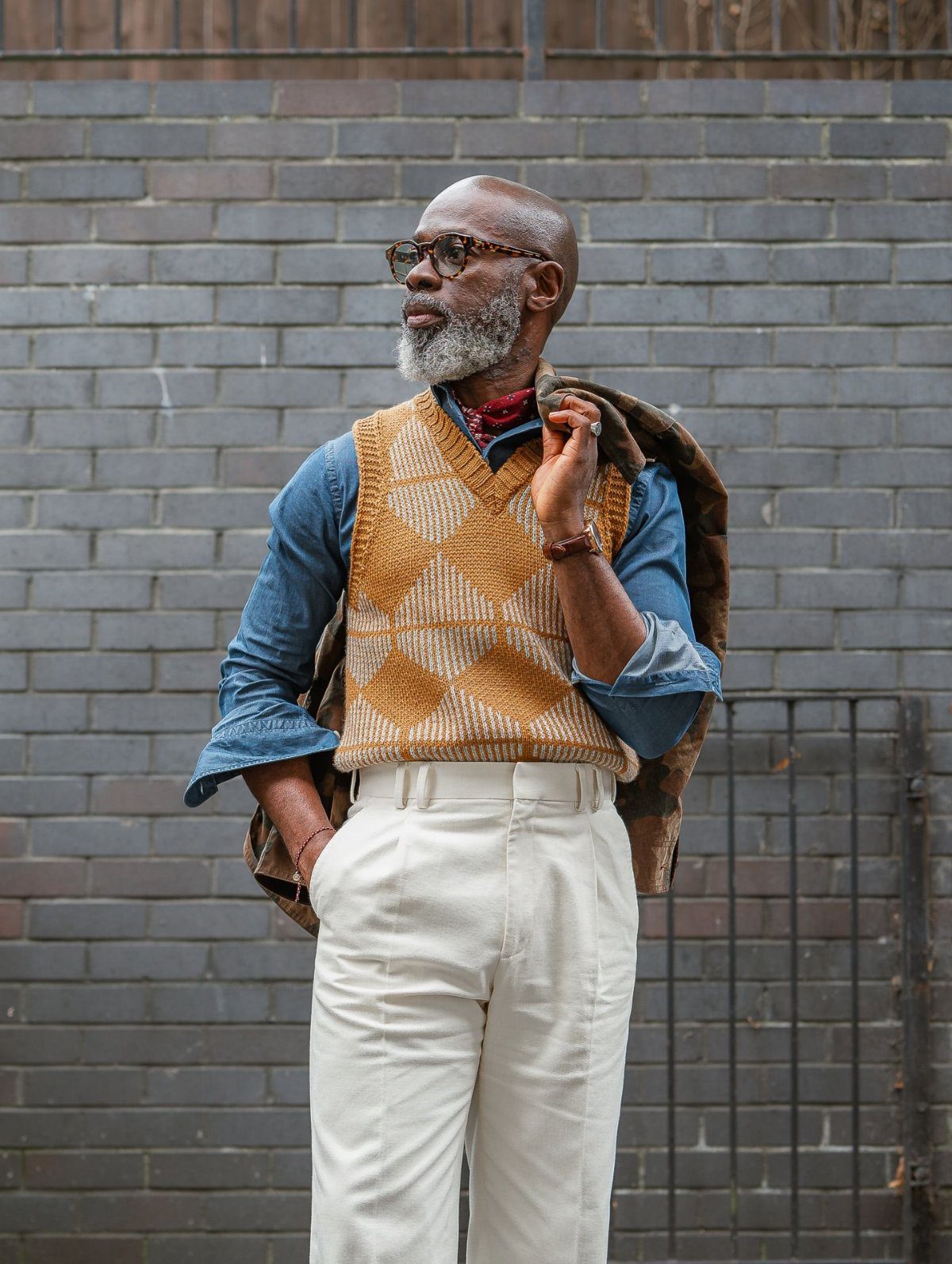 A man with a grey beard wearing a denim shirt, patterned sweater vest, and white trousers.
