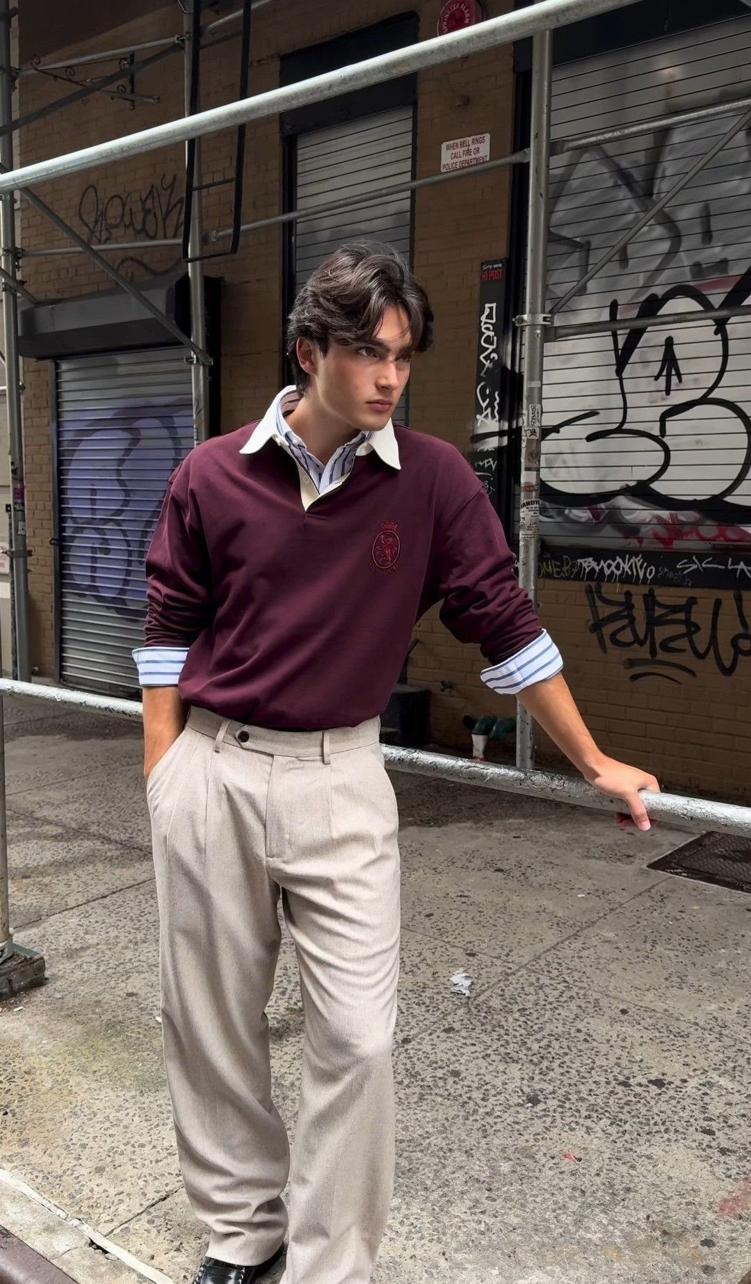 A young man wearing a maroon polo over a striped shirt and beige trousers.