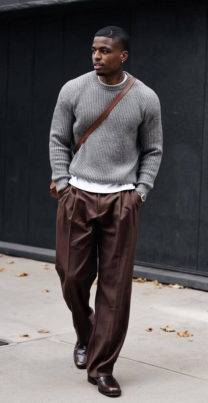 A man walking in a grey knit sweater, white t-shirt, and brown trousers.