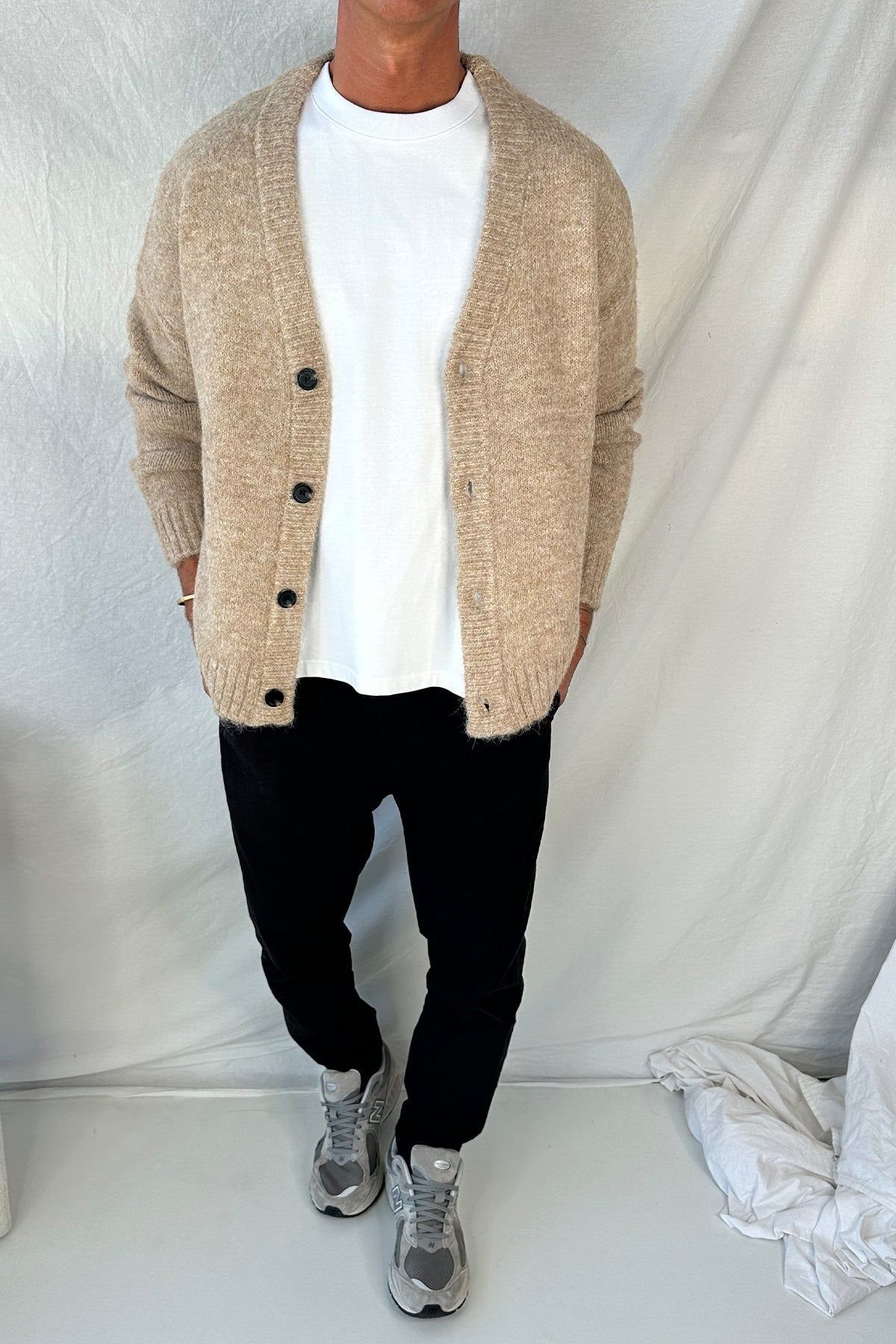 A person wearing a white t-shirt, camel cardigan, black trousers, and grey sneakers.