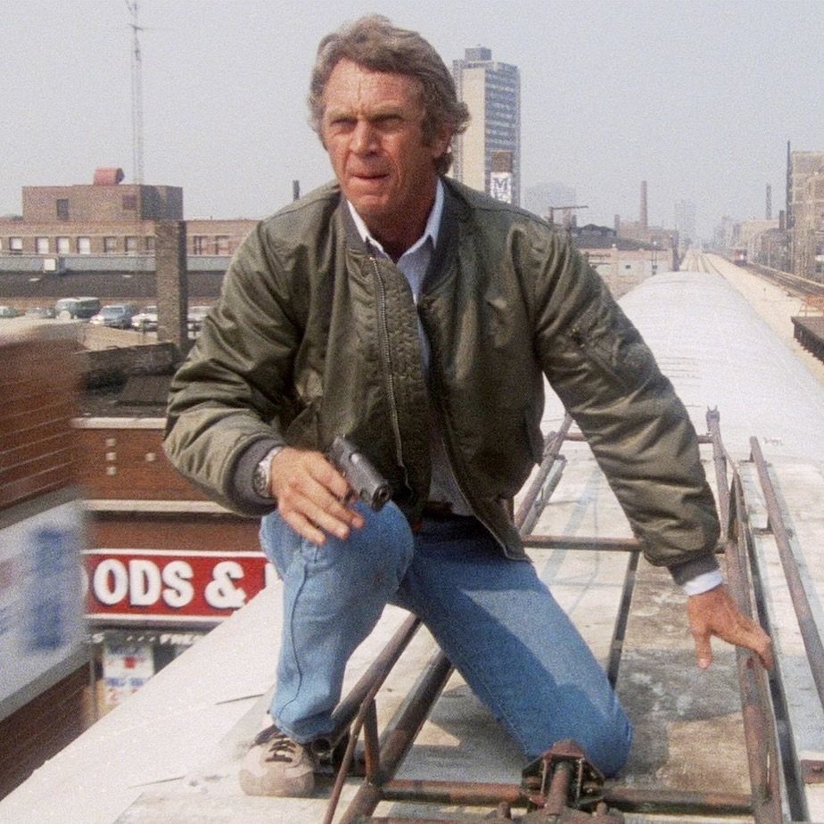 Man in a green bomber jacket and blue jeans holding a handgun on a rooftop.