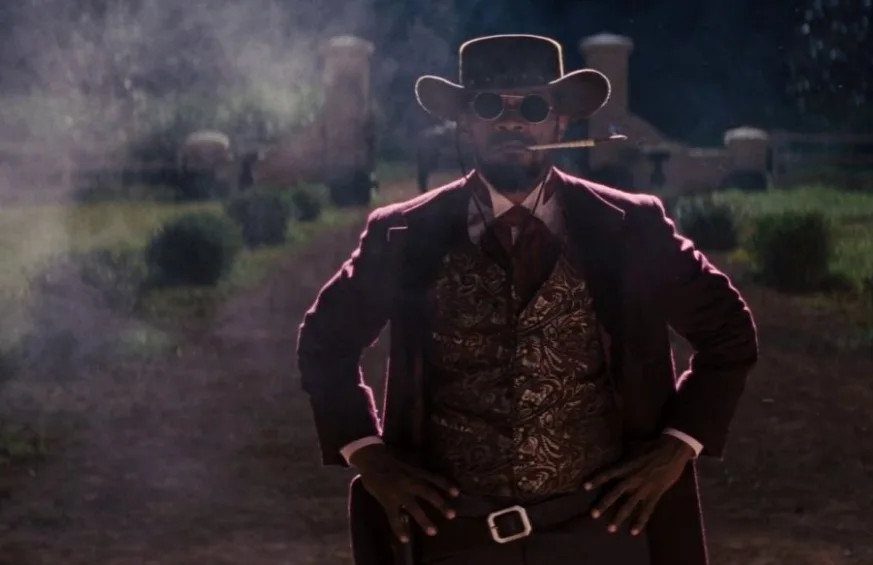 A man in a cowboy hat, sunglasses, and patterned waistcoat stands with hands on hips.